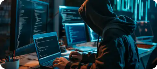 A hooded figure typing on a laptop in front of screens full of code